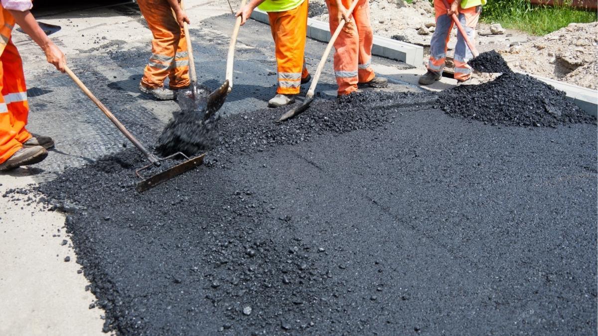 Comparing concrete vs asphalt driveways — a crew hand-raking fresh asphalt into place next to a concrete curb and base